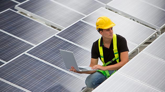 Männlicher Techniker kniet zwischen Solarpanelen mt einem Laptop in der Hand und testet die Anlage.