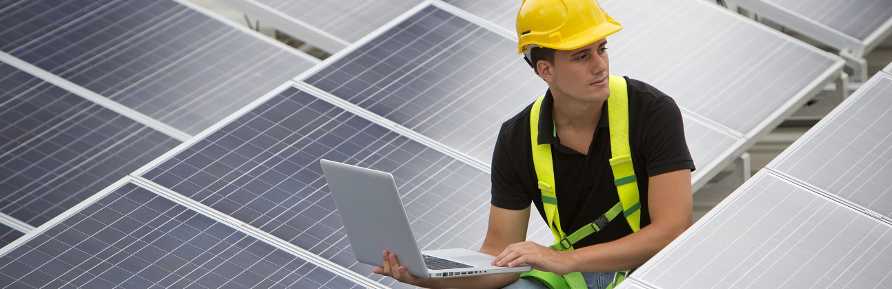 Das Bild zeigt einen Techniker mit Schutzhelm und Laptop der zwischen Solarpanelen kniet und die Anlage wartet.