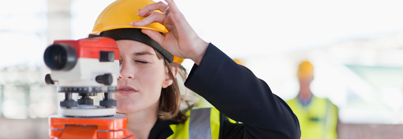 Vermessungstechnikerin mit gelbem Bauhelm blickt durch ein Vermessungsgerät. Das Bild symbolisiert Bau- und Infrastrukturprojekte die mit Mitteln des Europäischen Fonds für regionale Entwicklung (Ziel IWB) kofinanziert werden können.