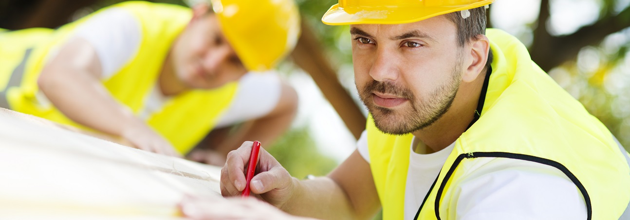 Zwei Zimmerer in Schutzausrüstung bearbeiten auf einer Baustelle Holzbalken.