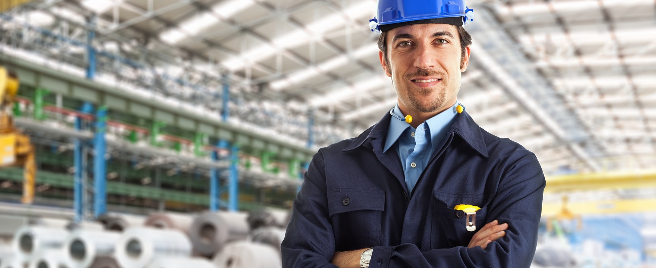 Mann mit Schutzhelm steht lächelnd und mit verschränkten Armen in der Mitte einer großen Lagerhalle, im Hintergrund sieht man Rollen mit Stahldraht.