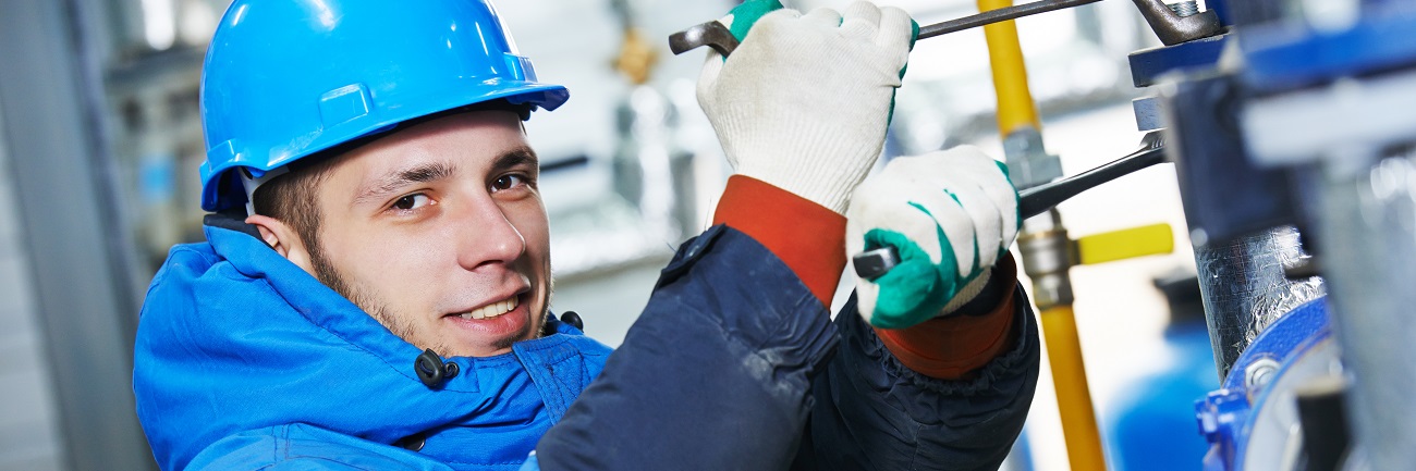Das Bild zeigt einen Mechaniker in blauer Arbeistuniform, der in jeder Hand einen Schraubenschlüssel hält und damit Schrauben einer modernen Maschine festzieht.