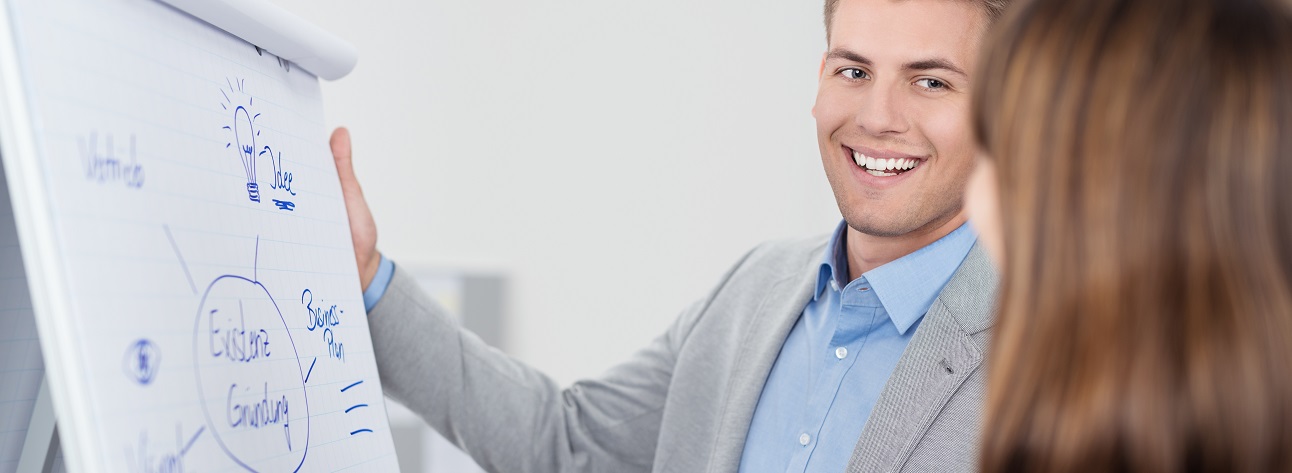 Mann und Frau stehen vor einem Flip-Chart, der Mann erläutert die darauf abgebildeten Eckpfeiler einer Unternehmensgründung.